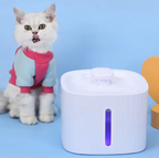 A cat wearing a pink sweater next to a white, wireless, automatic cat water fountain on a blue background